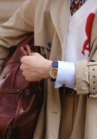 Abrigo trenzado color beige sobre una camisa azul claro con un suéter blanco debajo. El reloj de pulsera tiene una esfera beige y una correa azul; se sostiene un bolso de piel marrón.