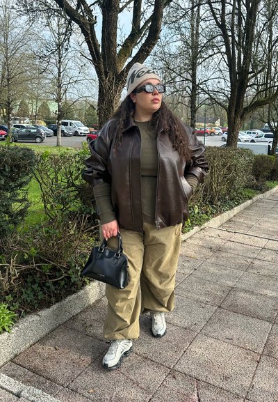 Persona con gafas de sol, un gorro, chaqueta de cuero oscura, suéter oliva, pantalones beige, zapatillas blancas, sosteniendo un bolso negro junto a un camino en el parque.