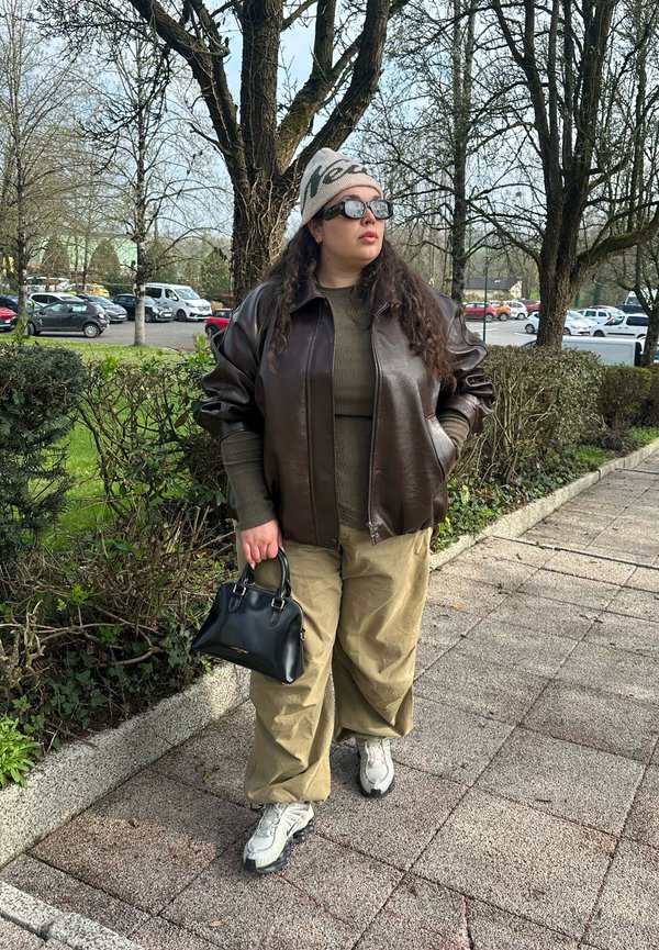 Persona con gafas de sol, un gorro, chaqueta de cuero oscura, suéter oliva, pantalones beige, zapatillas blancas, sosteniendo un bolso negro junto a un camino en el parque.