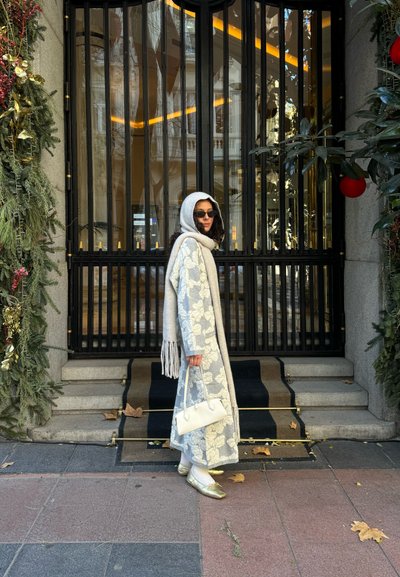 Abrigo largo gris con patrón floral blanco, bufanda gris, bolso blanco y bailarinas metálicas doradas. El fondo muestra puertas negras y vegetación.