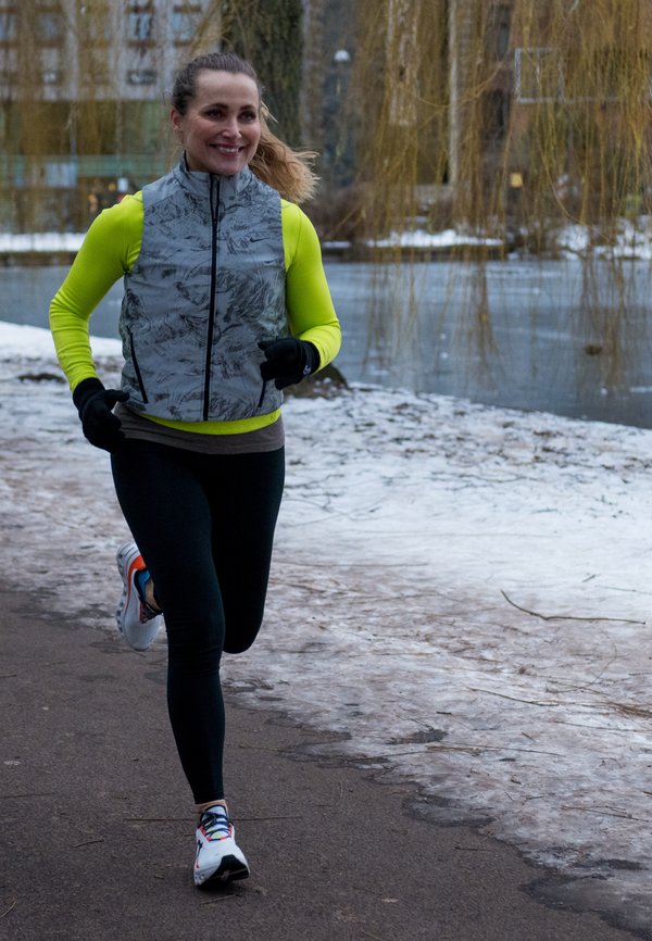 Woman jogging on a paved path beside a partially frozen pond, wearing bright yellow long-sleeved top, grey vest, black leggings, and white running shoes.