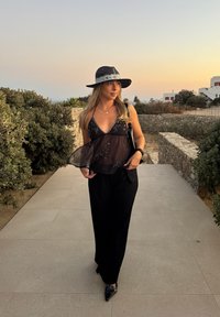 Črna prosojna majica s srebrnimi okraski, črne široke hlače, in črni klobuk s srebrnimi detajli. Zunanja postavitev s kamnito potjo.