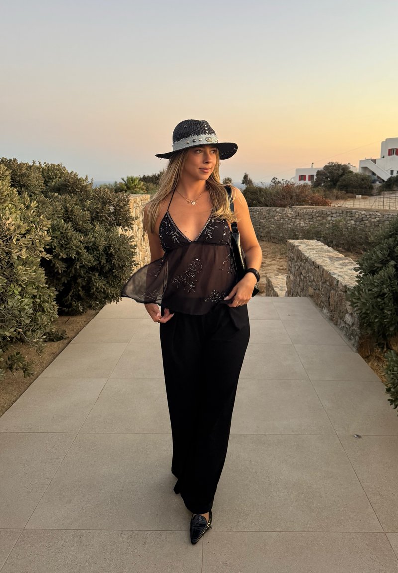 Haut noir transparent avec des embellissements argentés, pantalon noir à jambes larges, et chapeau noir avec des accents argentés. Cadre extérieur avec un chemin en pierre.