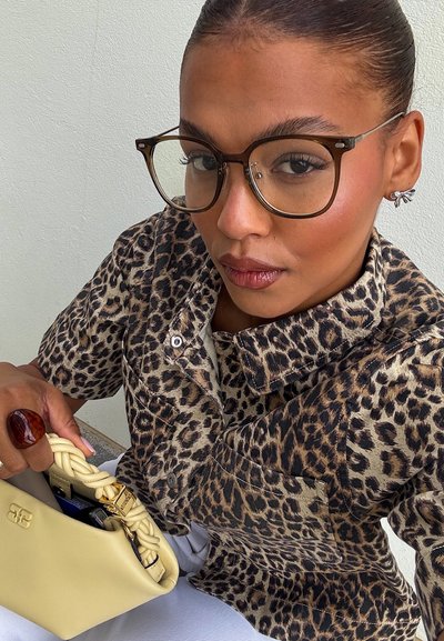 Chaqueta de estampado de leopardo con detalles de botones, anillo de ámbar y un bolso de mano crema con asa trenzada. Gafas con un marco delgado. Pantalones blancos visibles.