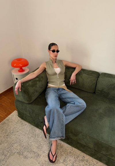 Mujer con gafas de sol, chaleco informal y vaqueros anchos descansando en un sofá verde de pana junto a una lámpara roja con forma de hongo sobre una mesita pequeña.