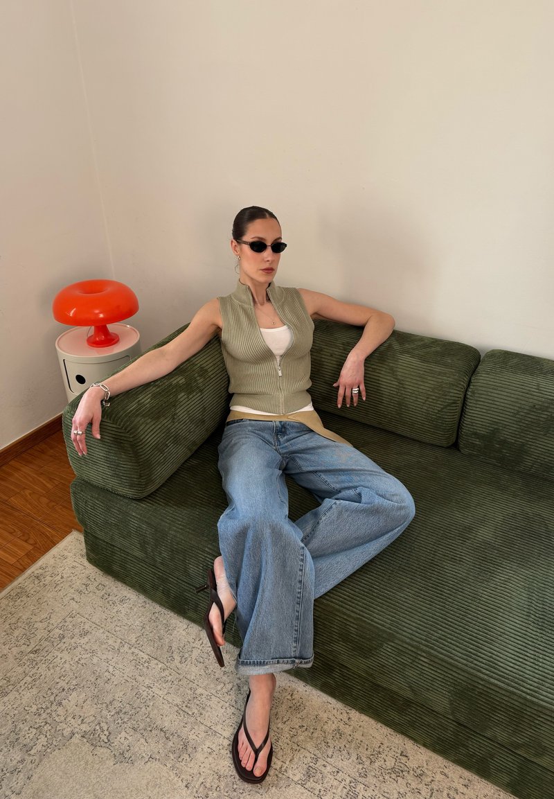 Femme portant des lunettes de soleil, un gilet décontracté et un jean large, allongée sur un canapé en velours côtelé vert à côté d'une lampe rouge en forme de champignon sur une petite table.