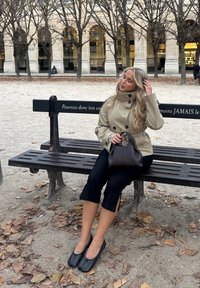 Beige trenssitakki, jossa napit yksityiskohtana, mustat lyhyet housut, mustat tasaiset kengät ja pieni ruskea käsilaukku, istumassa penkillä lehtien peittämänä.