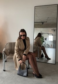 Femme en trench-coat beige, robe noire et talons assise sur une chaise, tenant un sac à main noir, reflétée dans un miroir en pied dans une pièce minimaliste.
