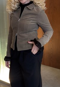 Hnědá manšestrová bunda se zipem vpředu, s žebrovanou texturou a černými průhlednými manžetami. Kombinovaná s volnými černými kalhotami, které představují minimalistický design.