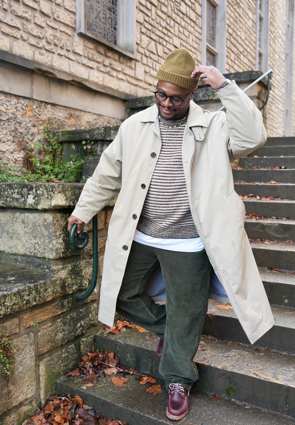 Beige waterproof coat, striped knitted jumper, green corduroy trousers, burgundy shoes. Stone steps with fallen leaves and greenery in the background.