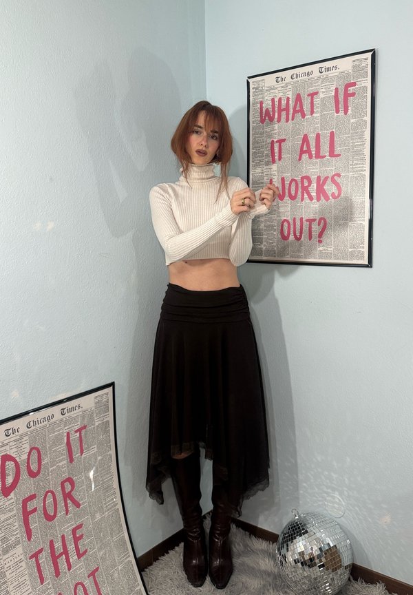 Mlajša ženska z rdečimi lasmi v belem kratkem puli in črni krilu pozira ob dveh uokvirjenih tiskih z roza besedilom in disko krogle na preprogi.