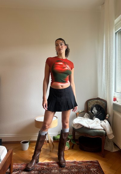 Top de manga corta en rojo y verde con un diseño estampado, combinado con una mini falda plisada negra y botas marrones hasta la rodilla con detalles en gris.