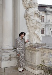 Personne portant un long manteau à motifs serpentins et un pantalon blanc, s'appuyant contre une statue en marbre détaillée sur une terrasse en pierre avec des bâtiments historiques.
