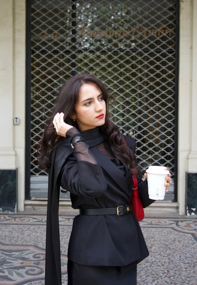 Mujer con ropa negra y mangas transparentes sosteniendo una taza de café blanca, ajustándose el cabello, parada frente a una puerta metálica cerrada.