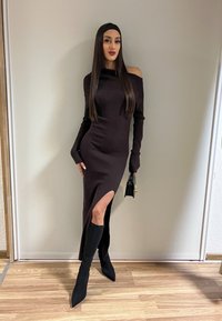 Femme aux longs cheveux raides porte une robe noire côtelée à épaules dénudées avec une fente latérale, des bottes noires montantes jusqu'aux genoux, et tient un petit sac à main noir.