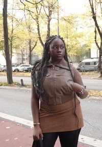 Veste en cuir marron, courte, avec fermeture éclair et ourlet côtelé. Associée à une jupe en daim marron. Cheveux longs, tressés et foncés, avec un bracelet en accessoire.