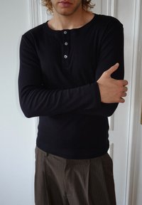 Maglia henley nera a maniche lunghe con tre bottoni argento sul collo, indossata con pantaloni marroni a righe sottili, che mostrano un tessuto liscio e un design aderente.