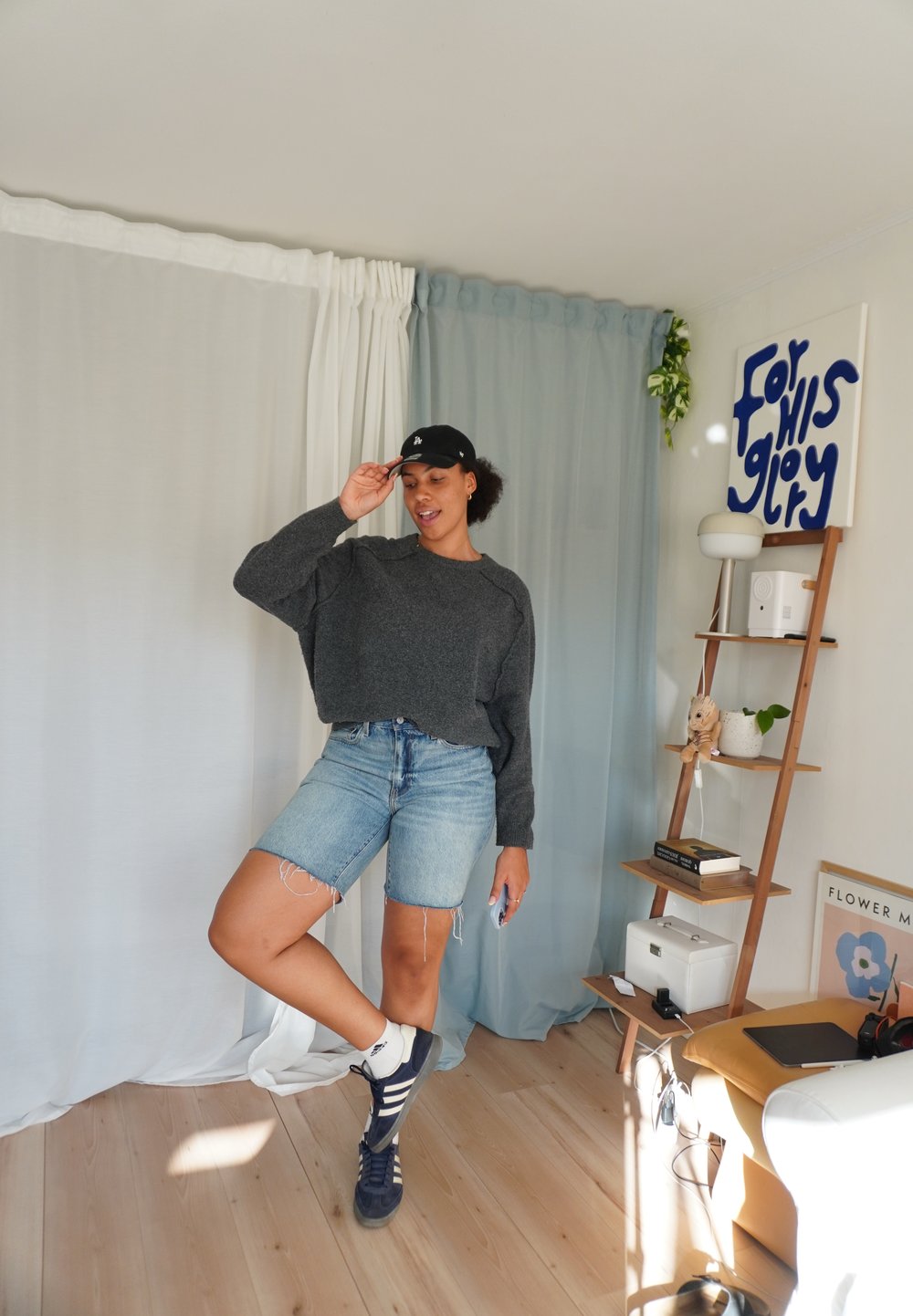 Sivkasta oversize pulover, modri jeans kratke hlače s frayed robovi, črna kapa in temni športni copati. Svetlo leseno tla in mehke zavese v ozadju.