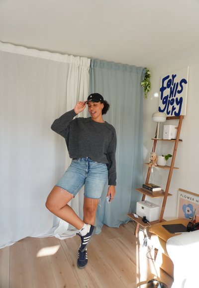 Suéter gris de gran tamaño, pantalones cortos de mezclilla azul con bordes deshilachados, gorra negra y zapatillas oscuras. Suelo de madera clara y cortinas suaves en el fondo.