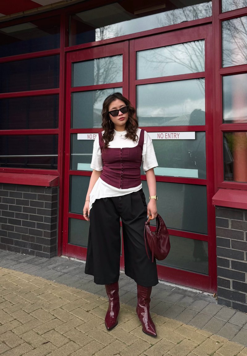 Femme portant des lunettes de soleil, un t-shirt blanc, un corset bordeaux, un pantalon noir à jambes larges, des bottes bordeaux, tenant un sac à main bordeaux assorti près de portes vitrées rouges.
