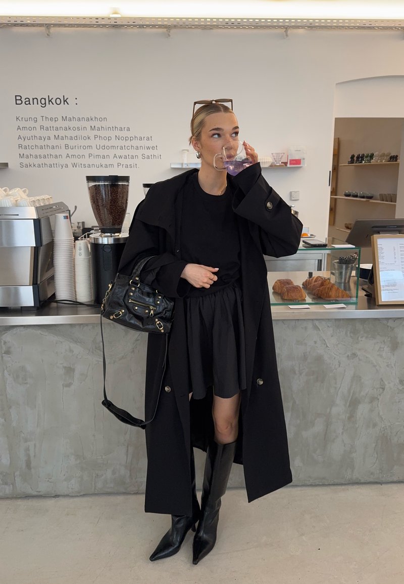Femme en manteau noir et bottes boit une boisson violette dans un café moderne près des pâtisseries et de la machine à café.