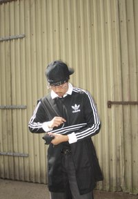 Juodas "Adidas" treniruotinis kostiumas su baltais dryžiais, baltu apykakle ir juoda kepure. Asmuo pritaiko savo rankovę, stovėdamas priešais šviesiai smėlinę gofruotą sieną.