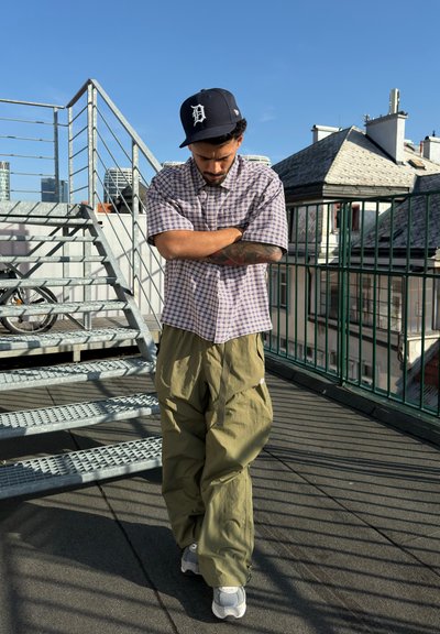 Camisa de manga corta a cuadros en blanco y marrón con pantalones holgados verdes; llevando una gorra negra. Configuración exterior en la azotea con escalera de metal.