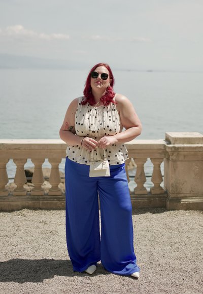 Blusa blanca con lunares negros y escote fruncido, combinada con pantalones anchos azules. Accesorios incluyen un pequeño bolso blanco. Fondo marítimo.