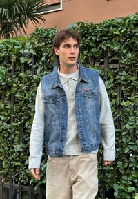 Farmerdzseki egy világosszürke pulóver felett, bézs kordbársony nadrággal párosítva. Természetes fényben, zöld növényzet háttérrel.