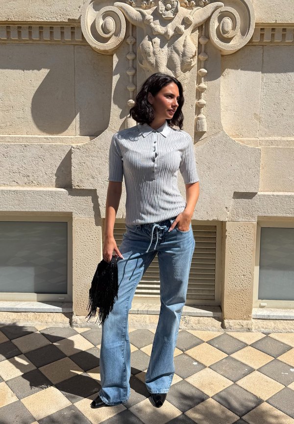 Femme debout contre un mur en pierre orné, portant un haut côtelé gris clair à manches courtes, un jean bleu, des chaussures noires pointues, et tenant un sac noir à franges.