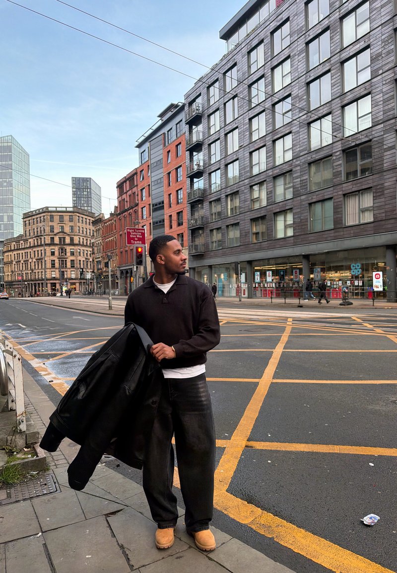 Un homme tenant une veste noire se tient sur un trottoir en ville près d'un passage piéton, avec des bâtiments modernes et historiques en arrière-plan.