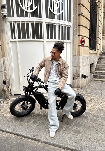 Bicicleta eléctrica negra con un diseño cuadrado, neumáticos gruesos y frenos de disco dobles, estacionada en una calle empedrada cerca de un edificio elegante.