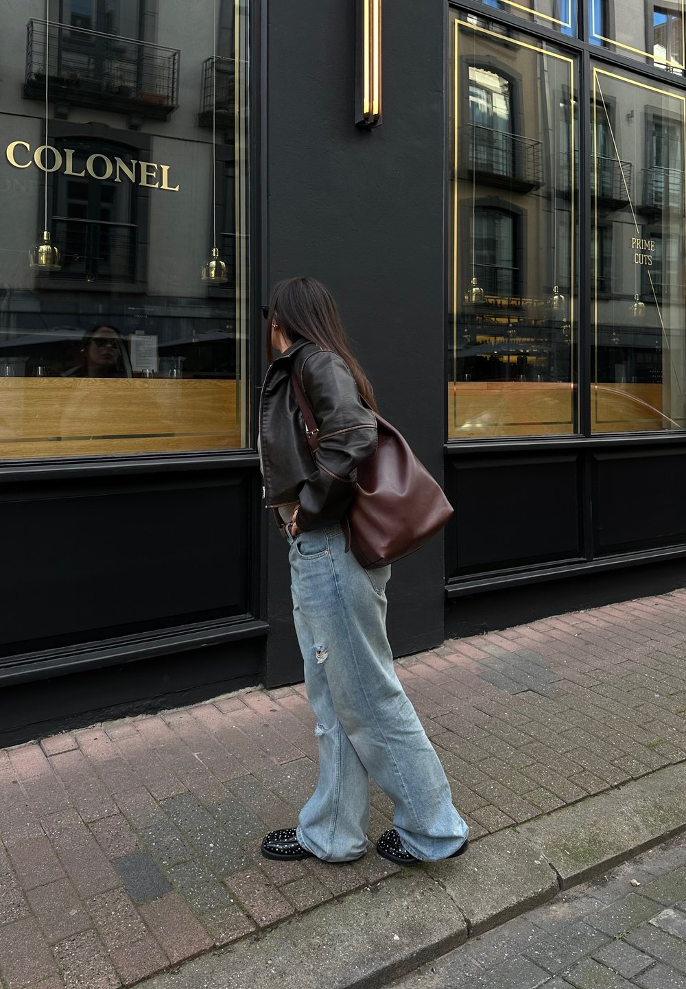 Pruun nahkjope, helesinised kulunud laiade jalgadega teksad, mustad studeeringutega kingad ja pruuni nahast käekott. Seisab restorani akna ligidal.