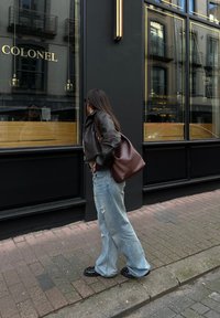 Pruun nahkjope, helesinised kulunud laiade jalgadega teksad, mustad studeeringutega kingad ja pruuni nahast käekott. Seisab restorani akna ligidal.