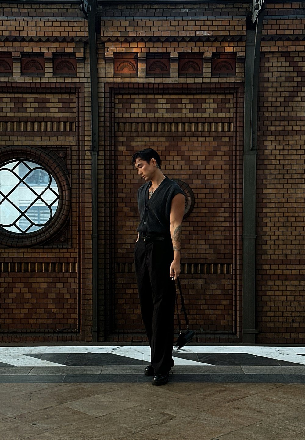 Black sleeveless button-up shirt, black trousers, holding a small bag. Standing against a textured brick wall with a round window.