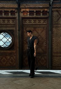 Black sleeveless button-up shirt, black trousers, holding a small bag. Standing against a textured brick wall with a round window.