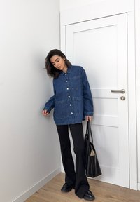 Modrá oversized košile z denimu s límcem, předními kapsami a patentními knoflíky, spárovaná s černými zvonovými kalhotami a texturovanou černou kabelkou.