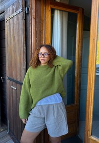 Grønn strikkegenser med vide ermer, lagd over en lyseblå skjorte. Kombinert med lysegrå shorts med tynne striper, stående ved en tredør.