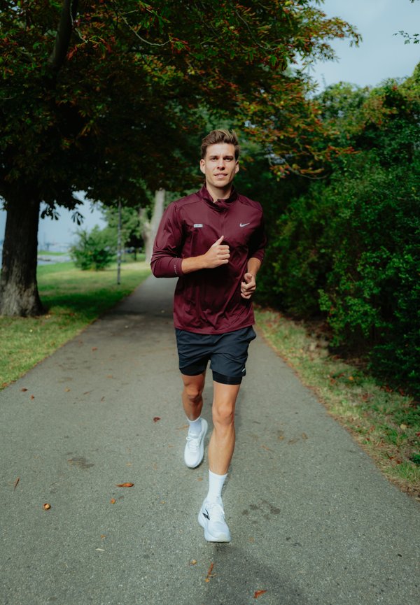 Chaqueta de running color burdeos con un cuello alto, pantalones cortos negros y zapatillas de running blancas. Corredor en un camino rodeado de árboles y vegetación.