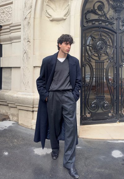 Joven con abrigo largo oscuro, suéter gris y pantalones anchos grises caminando por una calle de la ciudad cerca de un edificio de piedra tallada ornamentadamente y una puerta de hierro forjado negra.