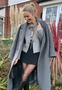 Γκρι μάλλινο παλτό, γκρι cropped πουλόβερ με λεπτομέρεια δέσμου, μαύρη μίνι φούστα, μαύρες ψηλές μπότες μέχρι το γόνατο, διαφανείς καλσόν με κουκίδες, πλεγμένη τσάντα χειρός.