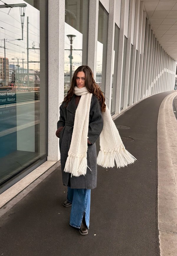 Žena s dugom smeđom kosom, u tamnom kaputu, širokim plavim trapericama, smeđim čizmama i dugim bijelim pletenim šal stojala na zakrivljenom pločniku.