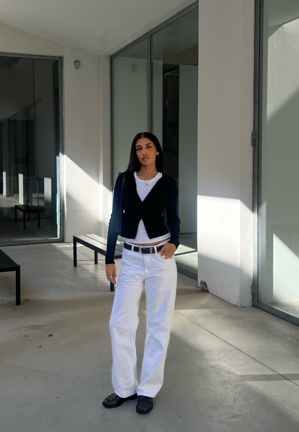 Cardigan corto nero sopra una maglietta bianca, abbinato a pantaloni bianchi larghi e scarpe scure. Una cintura e una collana argentate aggiungono tocchi di stile.