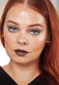 Close-up van een persoon met rood haar, blauwe ogen en metallic donkere lippenstift. Geaccentueerde jukbeenderen en subtiele glans oogschaduw in goudtinten.