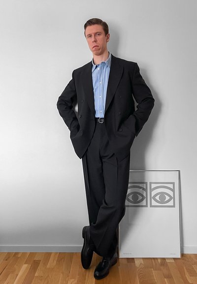 Hombre con traje negro y camisa azul a rayas, de pie con las manos en los bolsillos y las piernas cruzadas, contra una pared blanca con una obra de arte enmarcada de un ojo en el suelo.