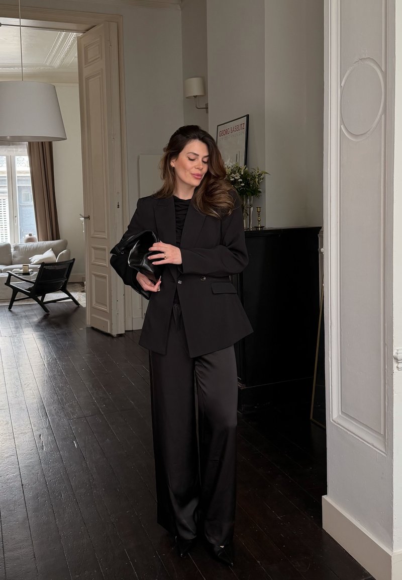 Woman in black pantsuit holding a matching clutch stands in a bright room with dark wooden floors and modern furniture.