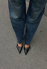 Sapatilhas pretas de bico fino com dois detalhes em fivela prateada, combinadas com jeans de ganga azul de corte largo, sobre um fundo de pavimento cinza.