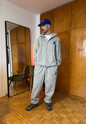 Hombre con chándal gris y gorra azul de pie en el interior cerca de una silla verde y un espejo alto, con una pared de madera como fondo.