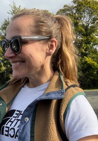 Chaleco de forro polar marrón con acentos en verde y azul marino sobre una camiseta blanca con texto negro. Gafas de sol negras, pequeños pendientes de aro naranjas.
