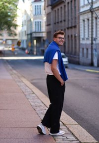 Fehér póló, kék pulóver a vállakon, fekete, széles szárú nadrág és fehér sportcipő; egy utcán áll, háttérben épületekkel.